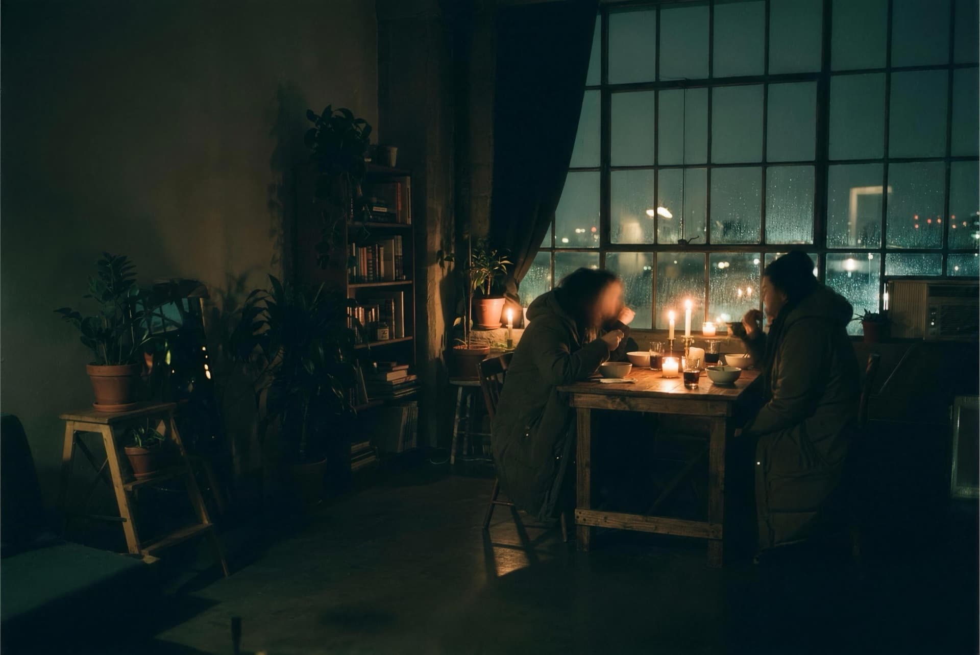 Two people share a quiet candlelit dinner by a rain-streaked loft window at night.